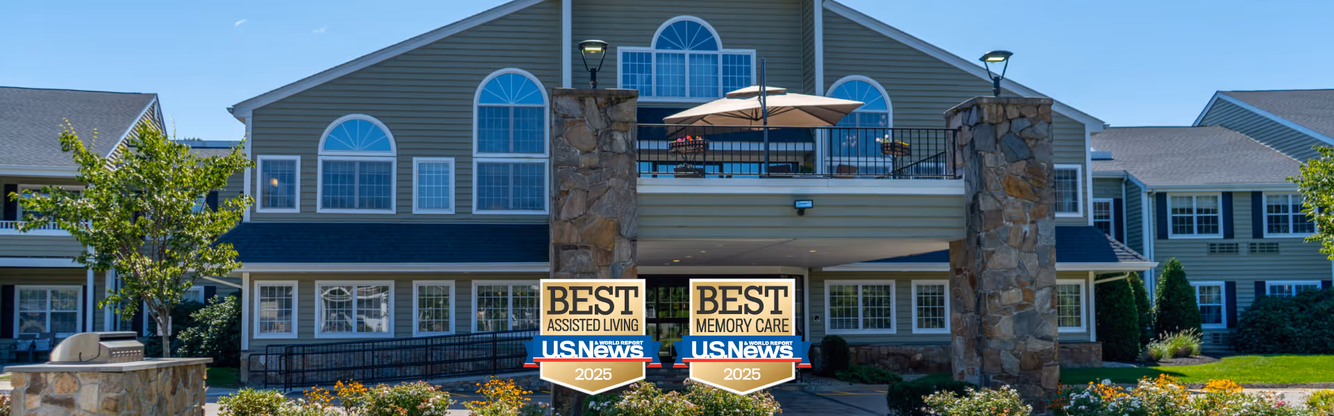 Front exterior view of a large two-story assisted living facility with green siding, multiple windows, stone pillars, and a balcony with outdoor seating. The building is surrounded by green grass, trees, and landscaping. Two signs in front display awards for Best Assisted Living and Best Memory Care by U.S. News for 2025.