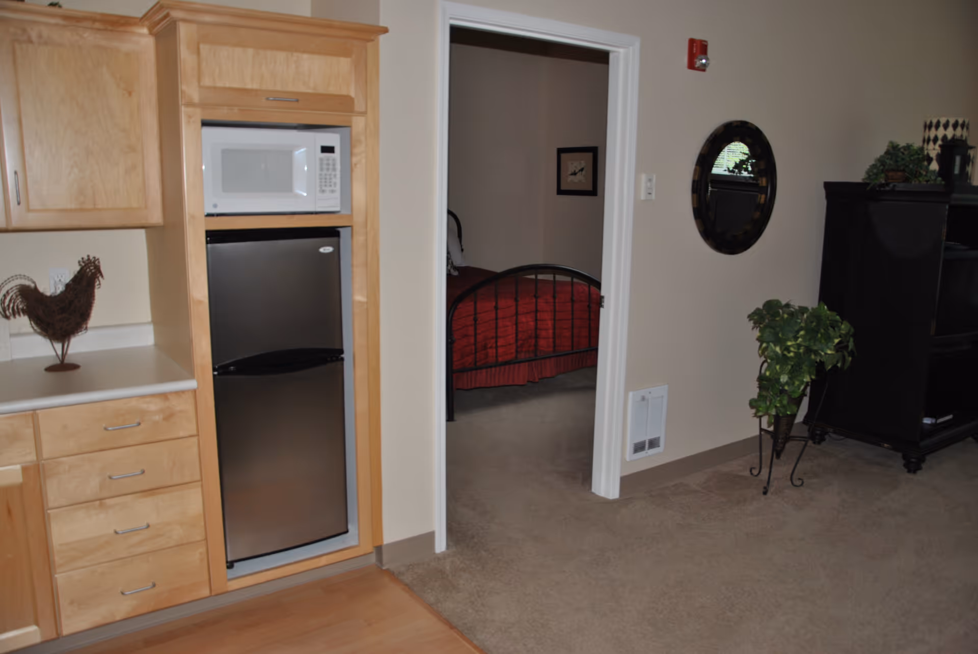 Interior view showing a small kitchen area with wooden cabinets, a microwave, and a stainless steel mini refrigerator. To the right, there is an open doorway leading to a bedroom with a bed covered in a red bedspread. The adjacent living area has a carpeted floor, a black entertainment cabinet, a round wall mirror, and a potted plant on a stand.