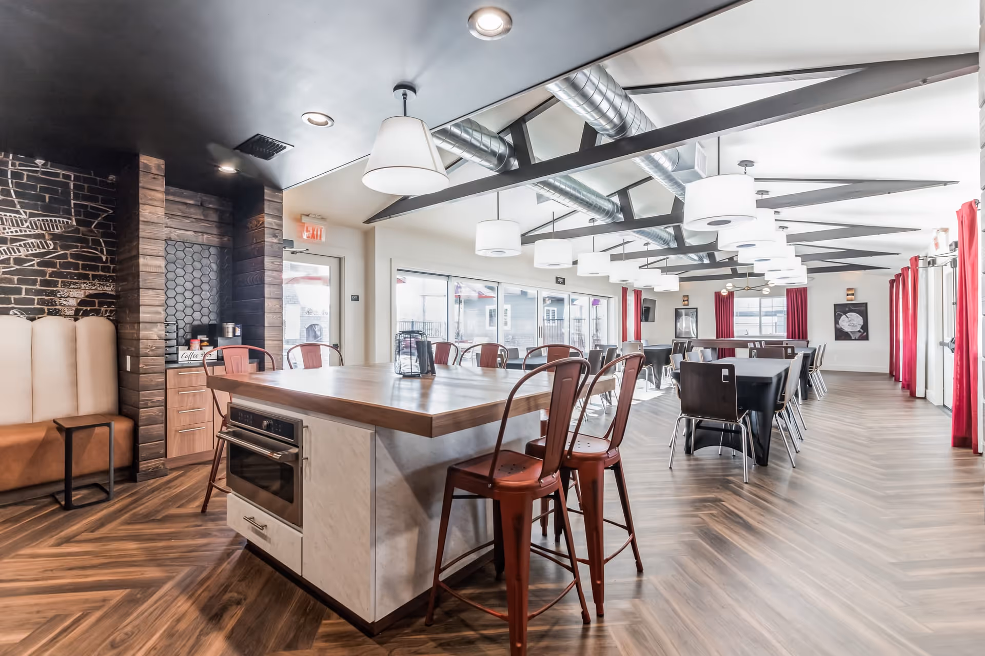 Spacious communal dining area with a central island, red metal bar stools, long dining tables, pendant lights and large windows with red curtains.