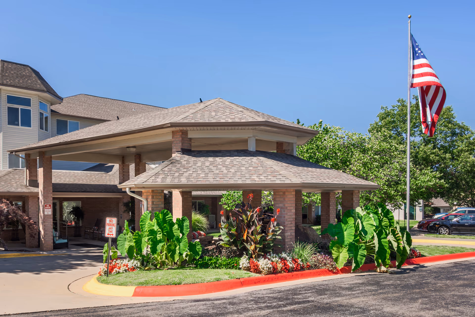 Entrance of The Gardens at Arkanshire Senior Living facility with a covered drop-off area, surrounded by green plants and flowers, an American flag on a flagpole, and a clear blue sky.