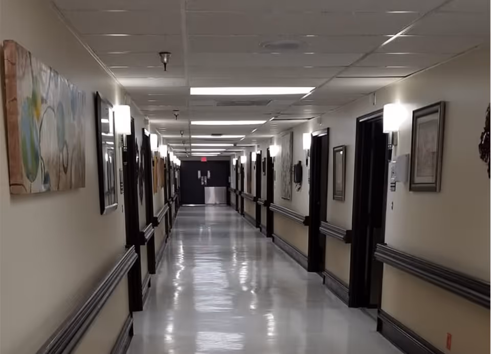 A long, clean hallway in a senior living facility with beige walls, dark wood trim, handrails on both sides, framed artwork hanging on the walls, and ceiling lights evenly spaced along the corridor.