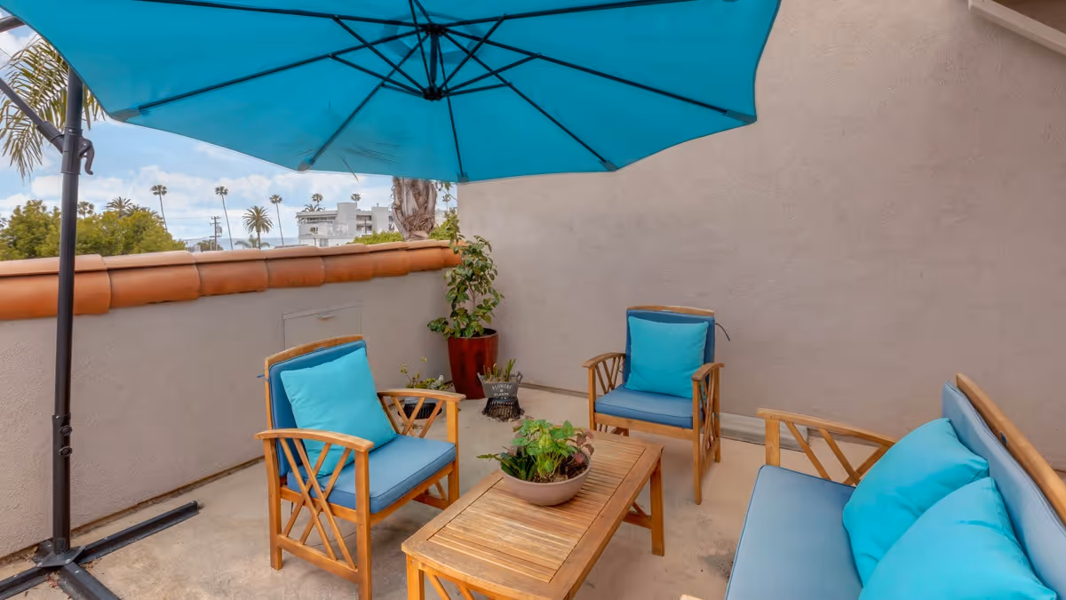 Outdoor patio area with wooden furniture including two armchairs and a loveseat, all with blue cushions. A wooden coffee table with a potted plant sits in the center. A large blue umbrella provides shade. The patio is enclosed by a beige wall with a terracotta tile top, and some greenery and palm trees are visible in the background.