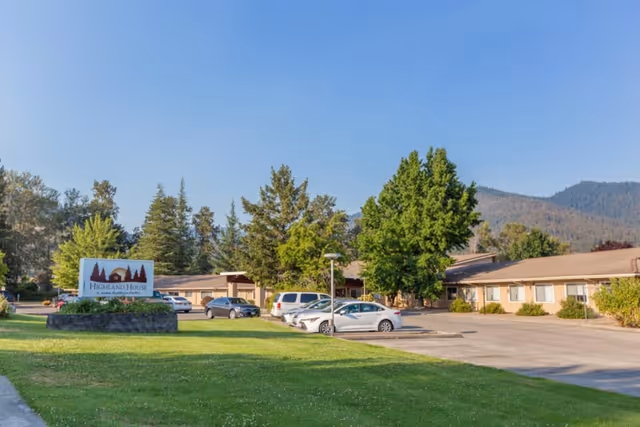 Front exterior of Highland House Nursing & Rehabilitation Center with its sign, parking lot with cars, trees, and distant hills.