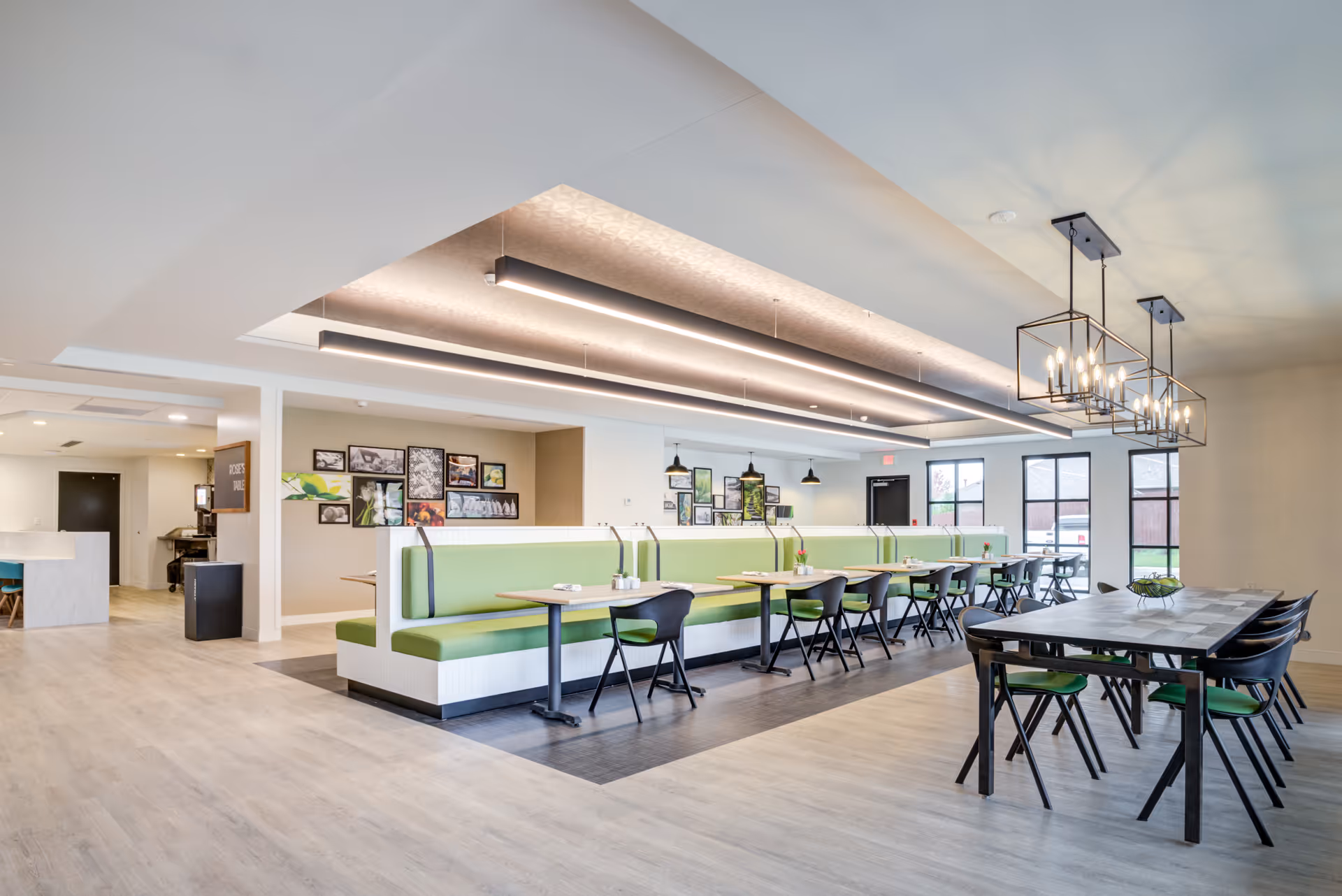 A bright and modern dining area with green cushioned booth seating along the wall and black chairs around wooden tables. The room features large windows letting in natural light, contemporary pendant lighting, and framed artwork on the walls. The flooring is light wood, and the space has a clean, open layout.