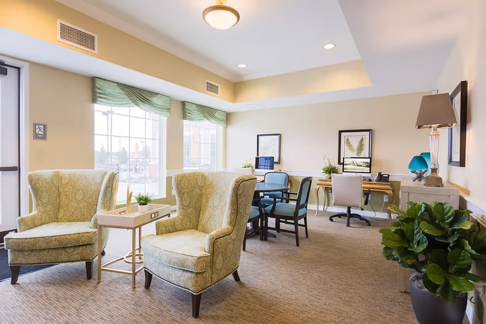 A bright and inviting common area with two patterned armchairs and a small table between them near large windows with green valances. In the background, there are desks with computers, chairs, framed botanical artwork on the walls, a table lamp, and a large potted plant on the right side.