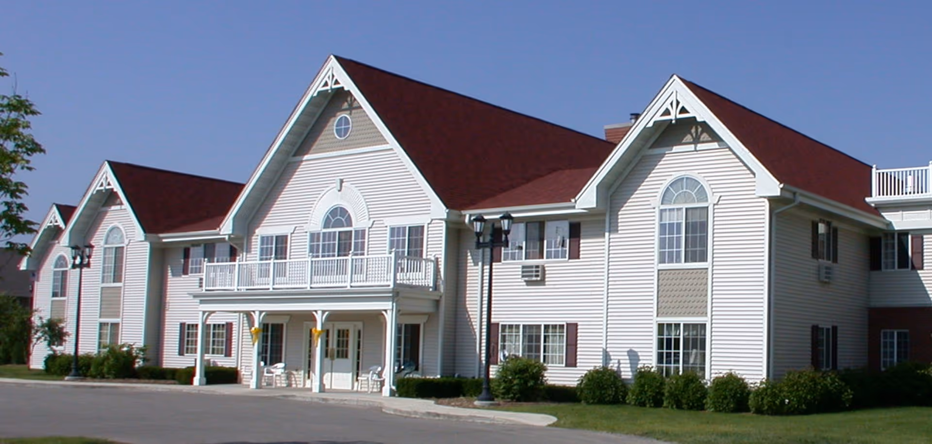 Front exterior of a two-story white senior living facility with red gabled roofs, balconies, and landscaped lawn.