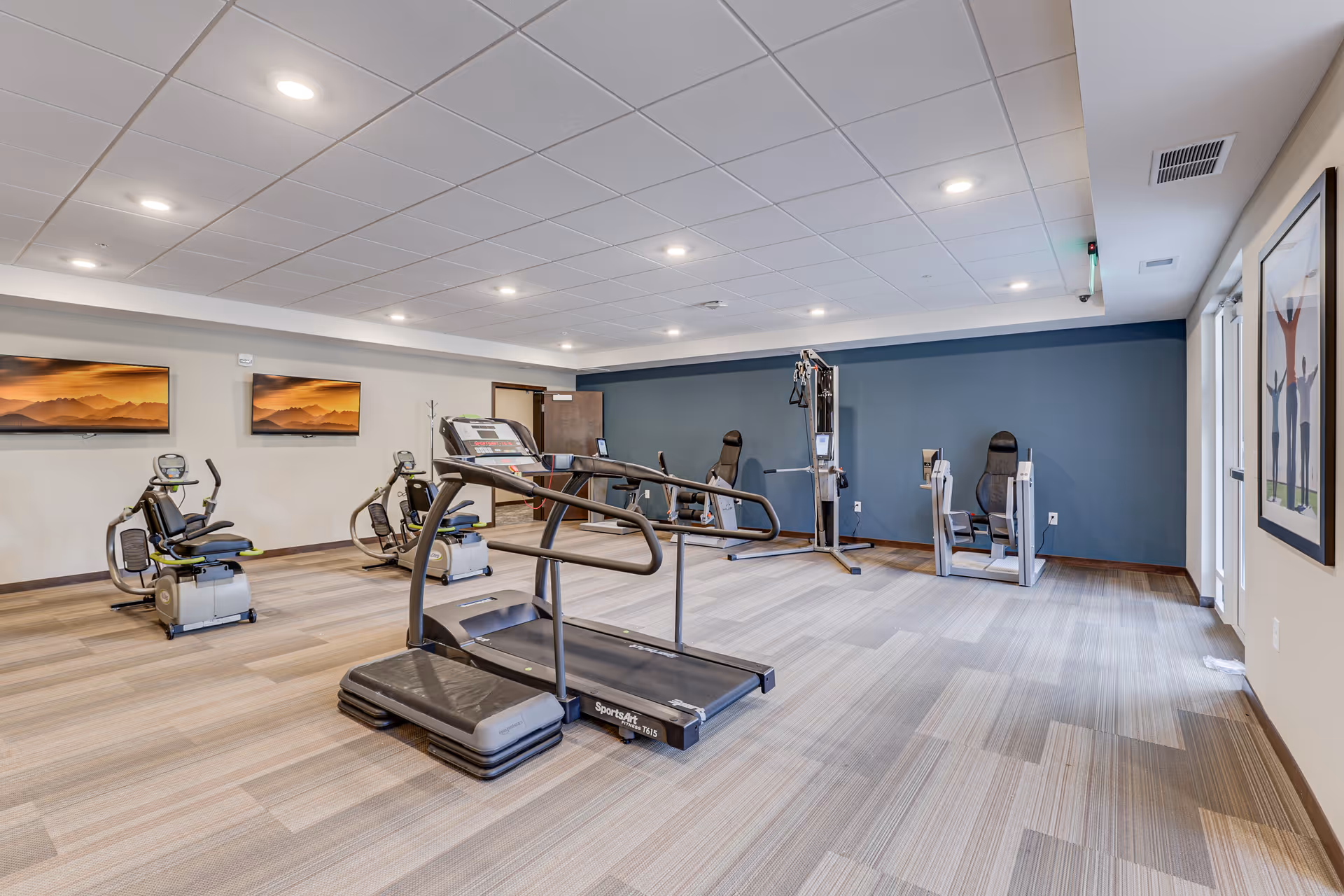 A fitness room in a senior living facility featuring exercise equipment including a treadmill, recumbent bikes, and strength training machines. The room has a light-colored carpet, a blue accent wall, ceiling lights, and two landscape paintings on the wall.
