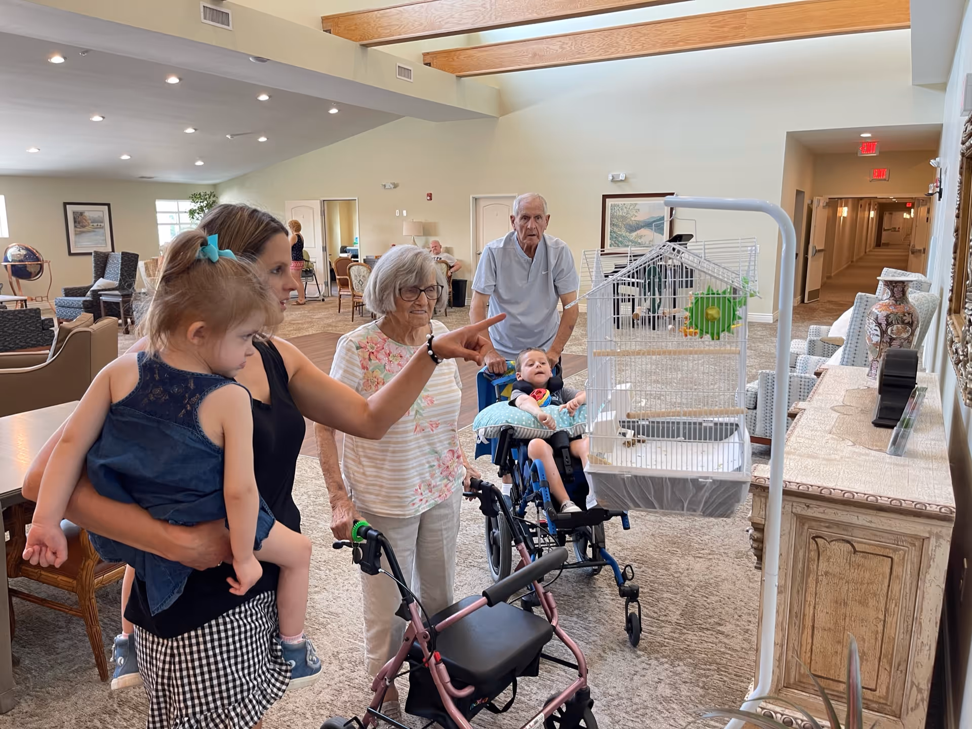 A group of people in a senior living facility common area. An elderly woman with a walker and an elderly man in a wheelchair are accompanied by a younger woman holding a small child. The younger woman is pointing towards a birdcage hanging on a stand. The room is spacious with high ceilings, wooden beams, and comfortable seating in the background.