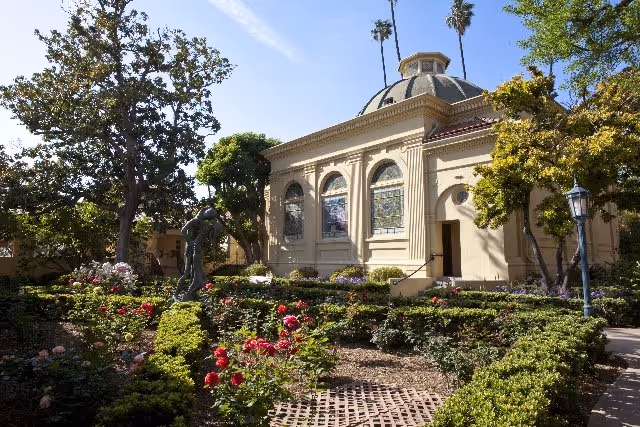 A sunny garden area with neatly trimmed bushes, blooming red and pink flowers, and a bronze statue of two figures. In the background, there is a beige building with arched windows and a domed roof, surrounded by tall palm trees and other greenery.