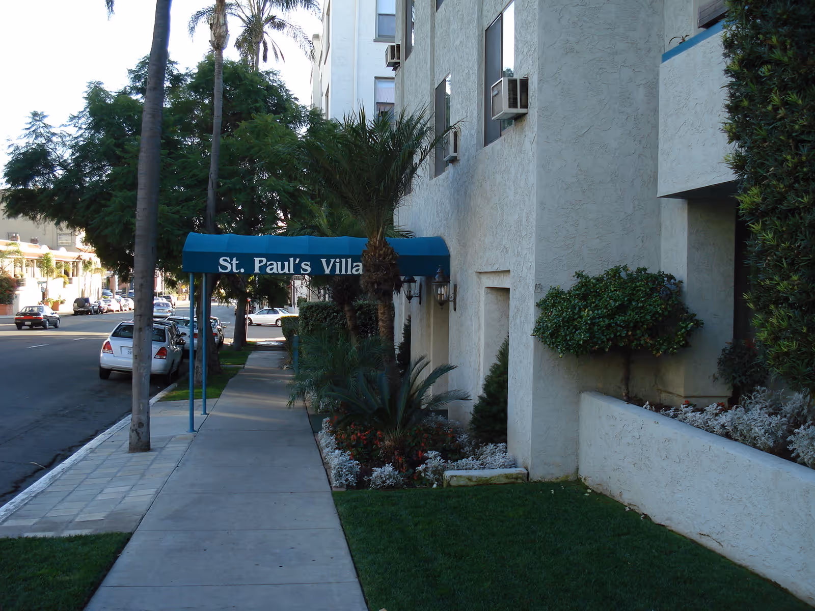Sidewalk view of a residential building with a blue awning that reads 'St. Paul's Villa'. The building has beige stucco walls, several windows with air conditioning units, and landscaped greenery including palm trees and bushes along the walkway.