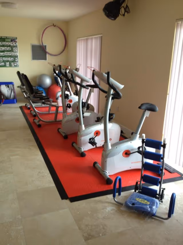 Indoor exercise room with three stationary bikes lined up on a red mat, two recumbent exercise bikes behind them, exercise balls, a hula hoop hanging on the wall, and an ab workout machine on the floor near the bikes. The room has beige tiled flooring and vertical blinds covering the windows.