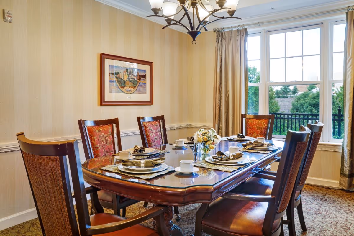 A formal dining room with a polished wooden table set for six people. The table has placemats, plates, cups, and napkins arranged neatly, with a small floral centerpiece in the middle. The room features beige striped wallpaper, a framed picture on the wall, a chandelier overhead, and large windows with beige curtains letting in natural light and showing greenery outside.