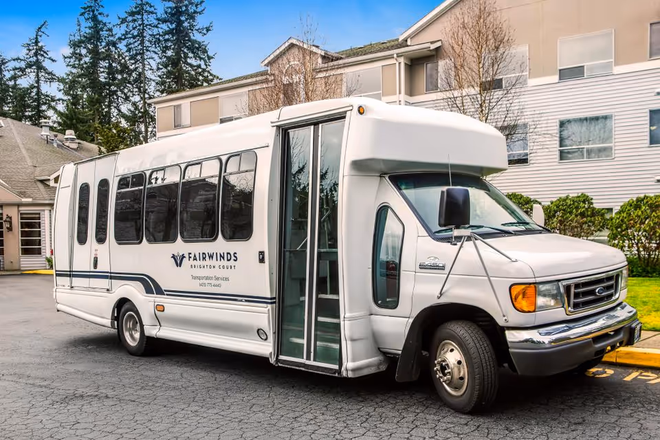 A white shuttle bus parked outside a building with the logo and text 'Fairwinds Brighton Court Transportation Services' on its side. The bus has large windows and an open door, with trees and a multi-story building in the background.