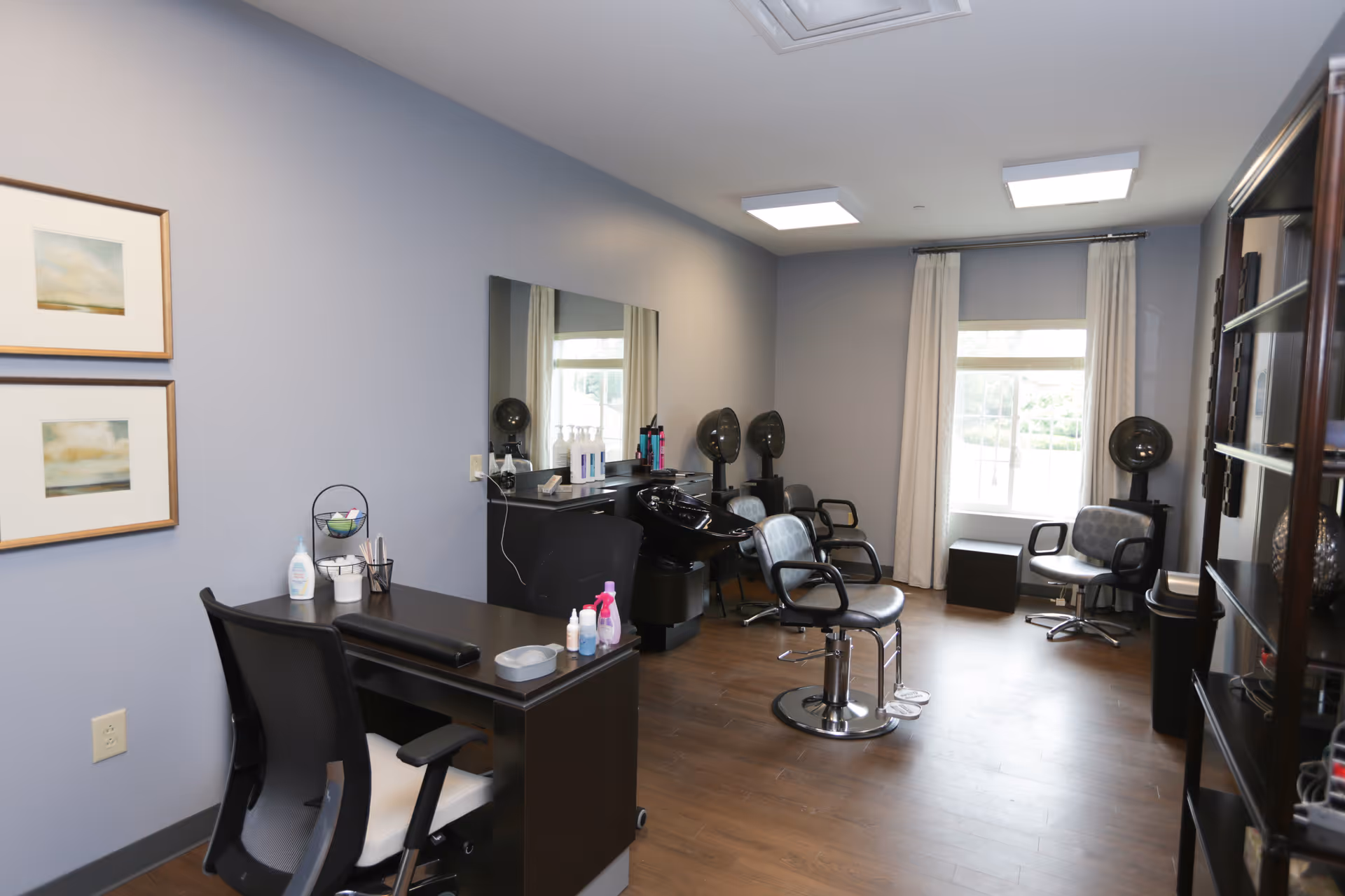 A well-lit salon room with a desk and chair in the foreground, salon chairs with hair drying stations along the wall, a large mirror, and a window with curtains letting in natural light. The room has wooden flooring and light gray walls with two framed pictures.
