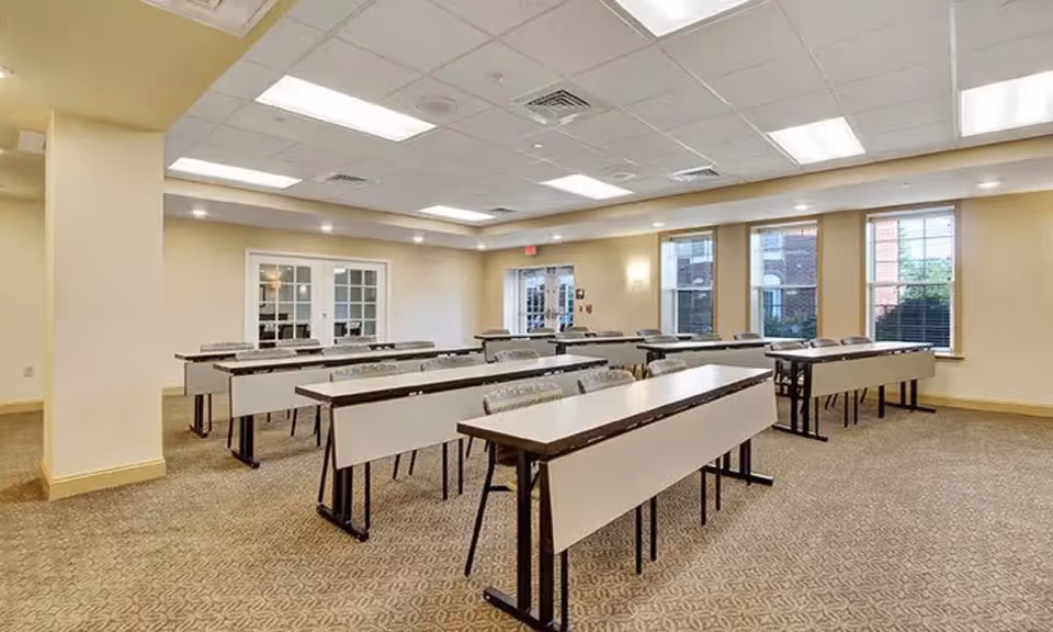 Bright activity/meeting room with rows of long tables and chairs and windows along one wall.