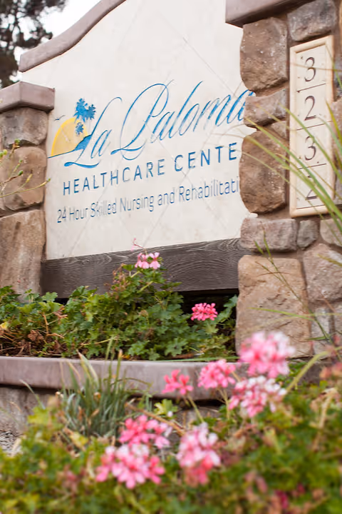 Stone sign for La Paloma Healthcare Center surrounded by green plants and pink flowers, with the address number 323 visible on the right side.