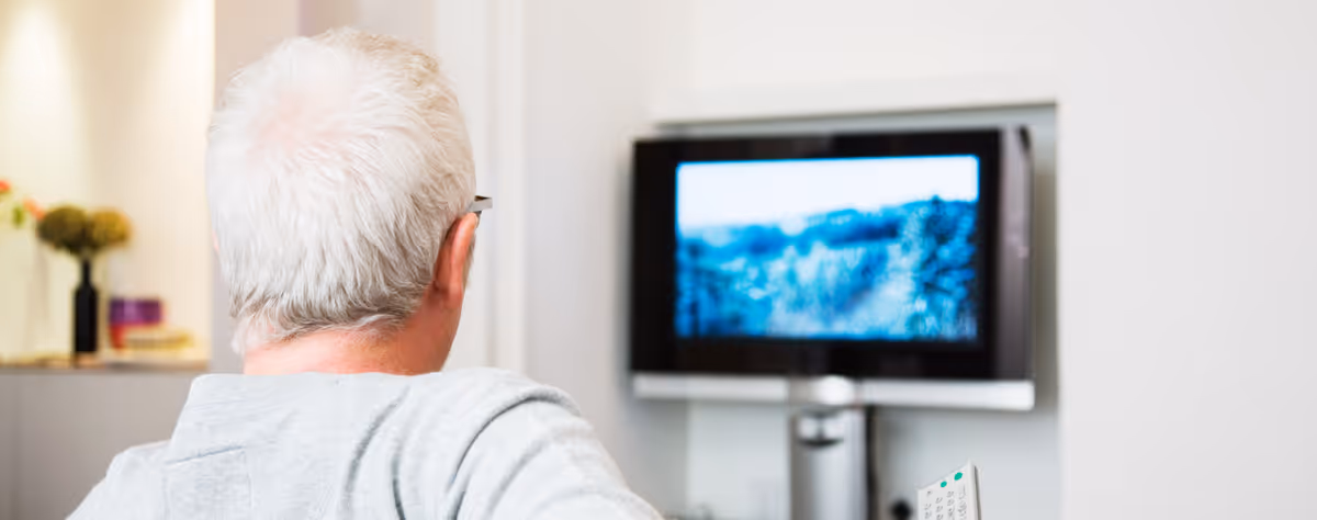 An elderly person with white hair sits in a living room watching a wall-mounted TV.