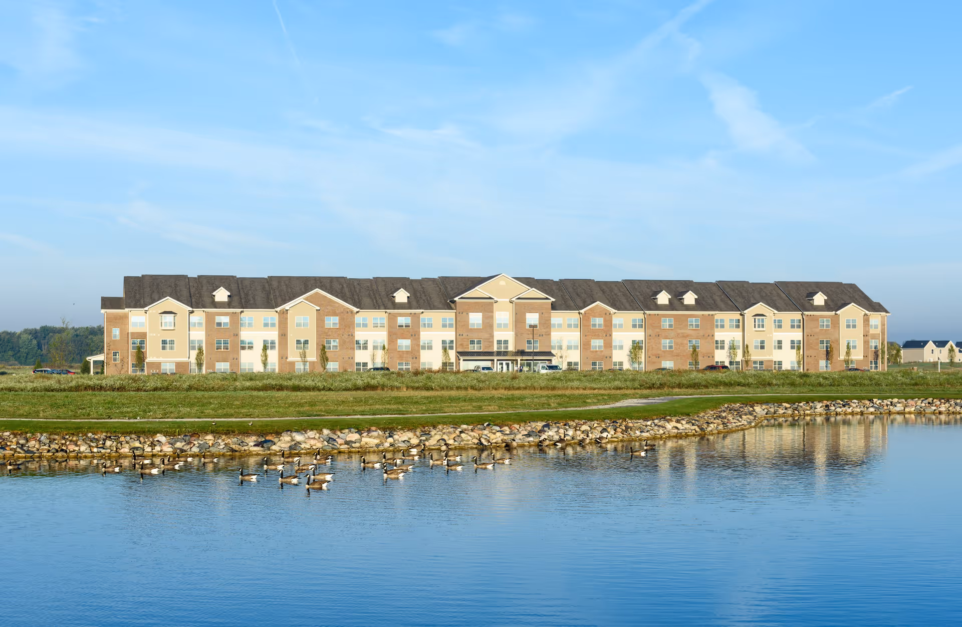 Wide three-story senior living building seen across a pond with geese under a blue sky.