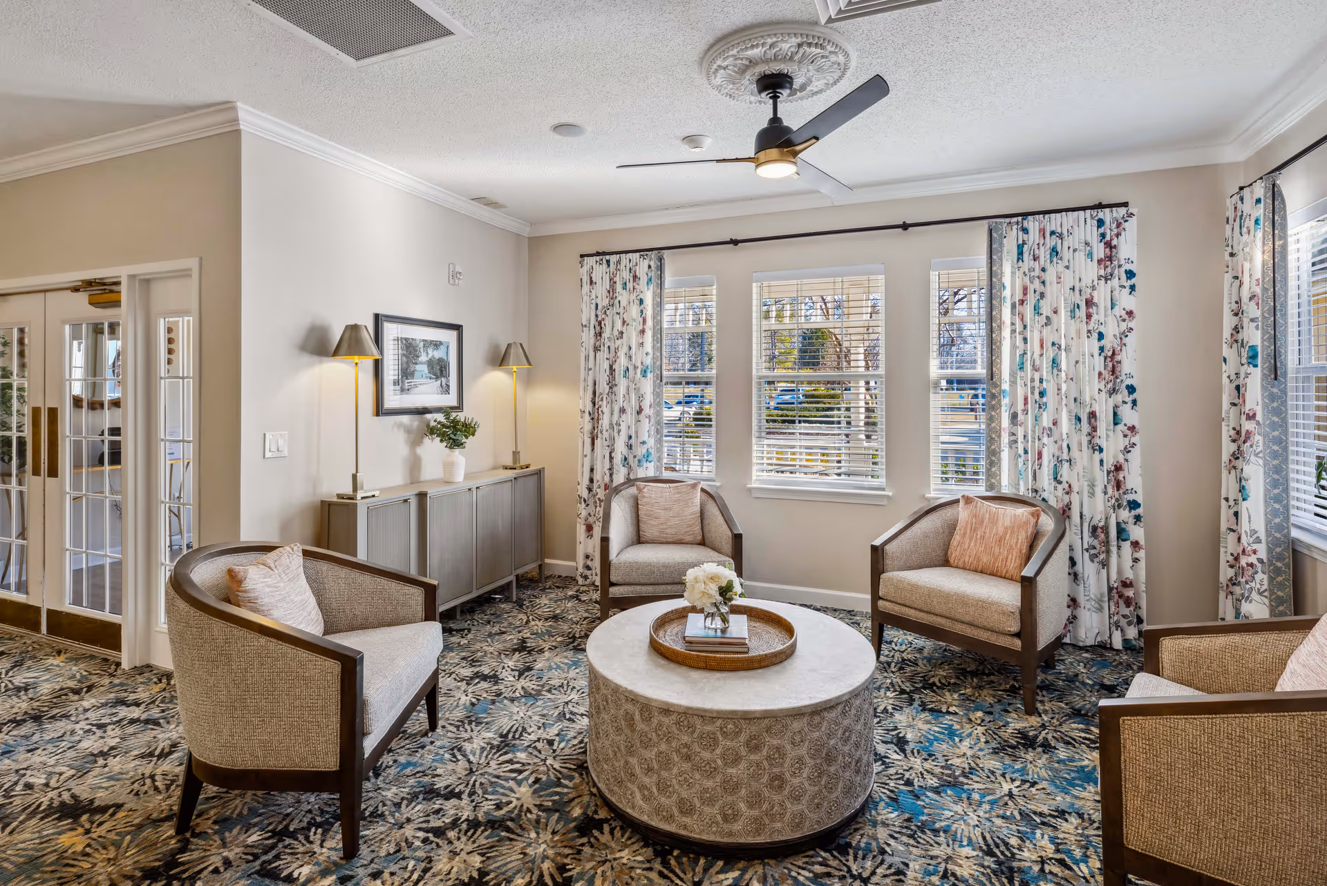 Bright furnished sitting room with four upholstered armchairs around a round coffee table, patterned carpet, and windows with floral curtains.