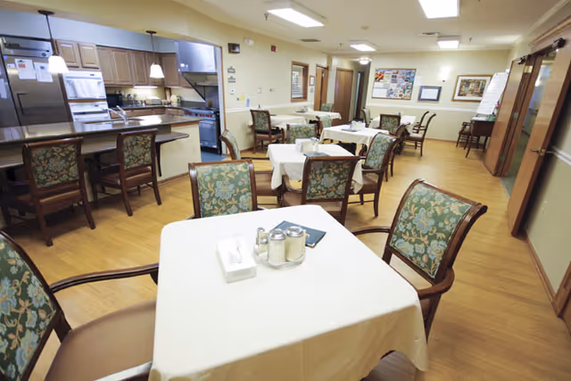 A dining room in a senior living facility with several tables covered with white tablecloths and surrounded by wooden chairs with floral upholstery. The room has a wooden floor and a kitchen area with appliances and a counter with stools in the background. The walls are light-colored with framed pictures and a bulletin board.
