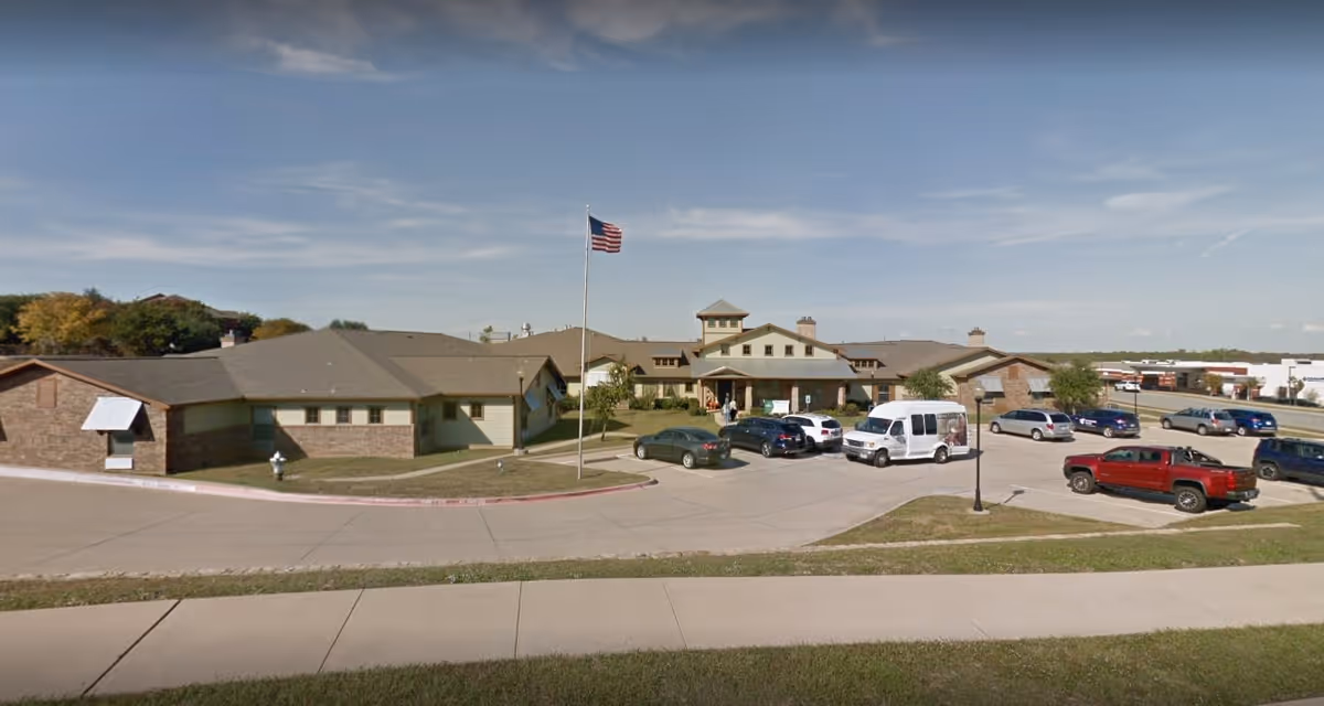 Exterior view of Pecan Point Assisted Living and Memory Care facility showing a single-story building with a central entrance, an American flag on a flagpole in front, several parked cars, and a clear sky.