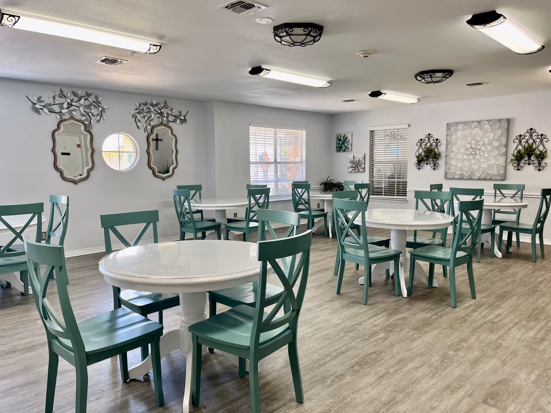 Dining room with round white tables and teal chairs on wood-look flooring and decorative wall art.