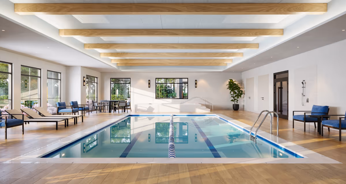 Indoor swimming pool area with wooden flooring and ceiling beams. The pool has a lane divider and metal handrails. Around the pool are lounge chairs and seating areas with blue cushions. Large windows let in natural light and show greenery outside. A potted plant and a wall-mounted shower are visible on the right side.