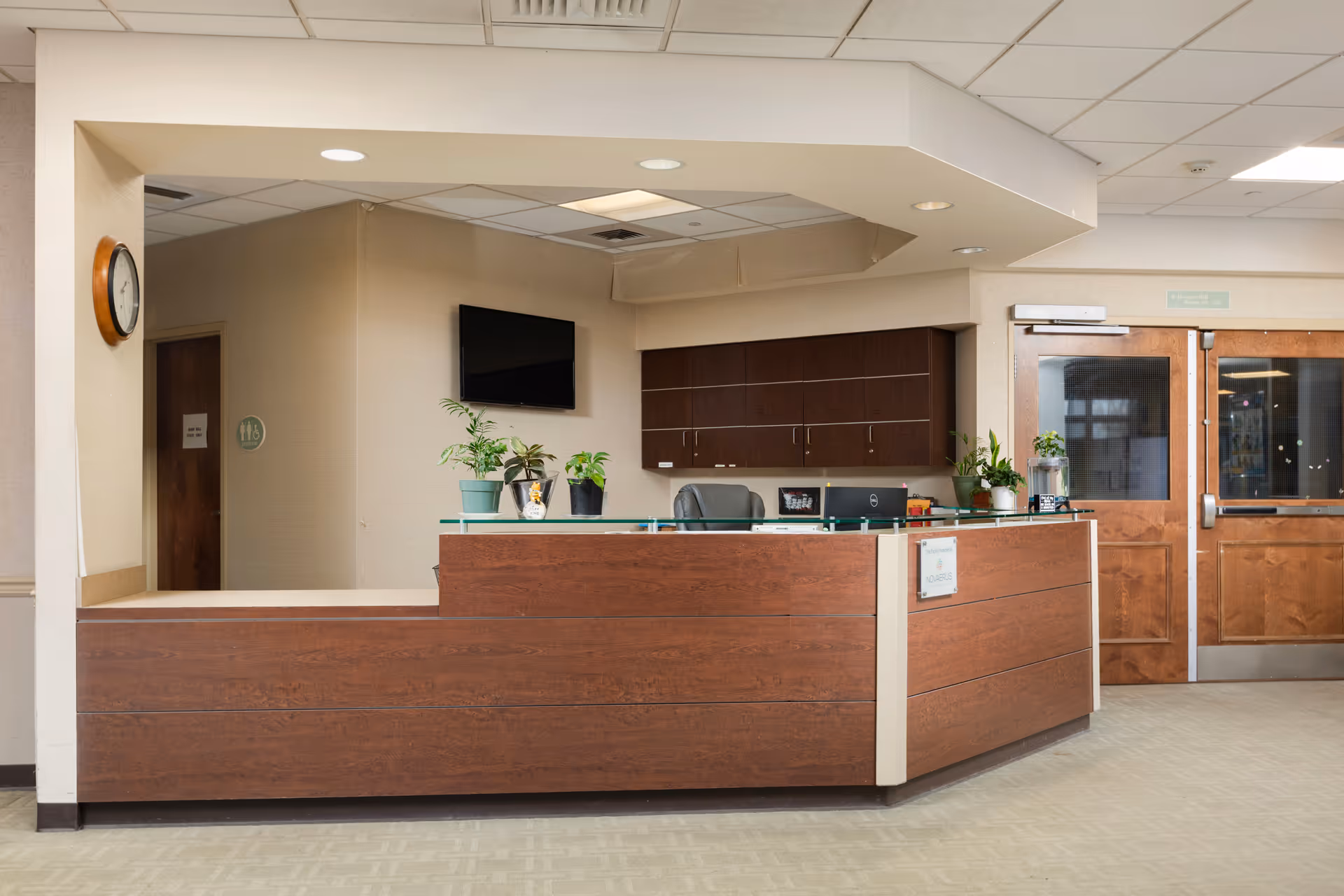 Reception desk area in a senior living facility with a wooden counter, several potted plants, a wall-mounted TV, wooden double doors, and a clock on the wall.