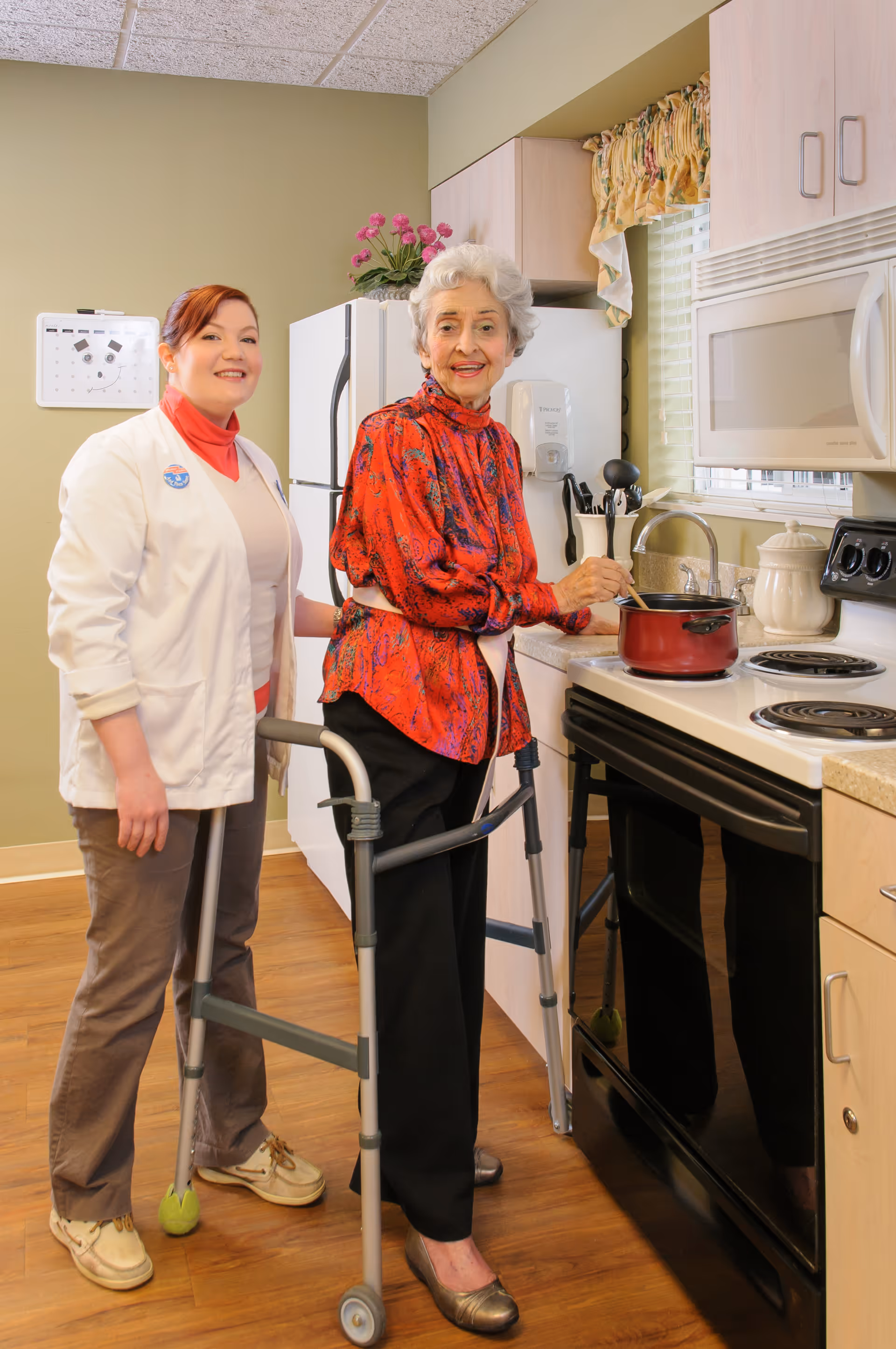 An elderly woman using a walker is stirring a pot on the stove in a kitchen, while a caregiver stands beside her smiling. The kitchen has light-colored cabinets, a microwave, a refrigerator, and a window with floral curtains.