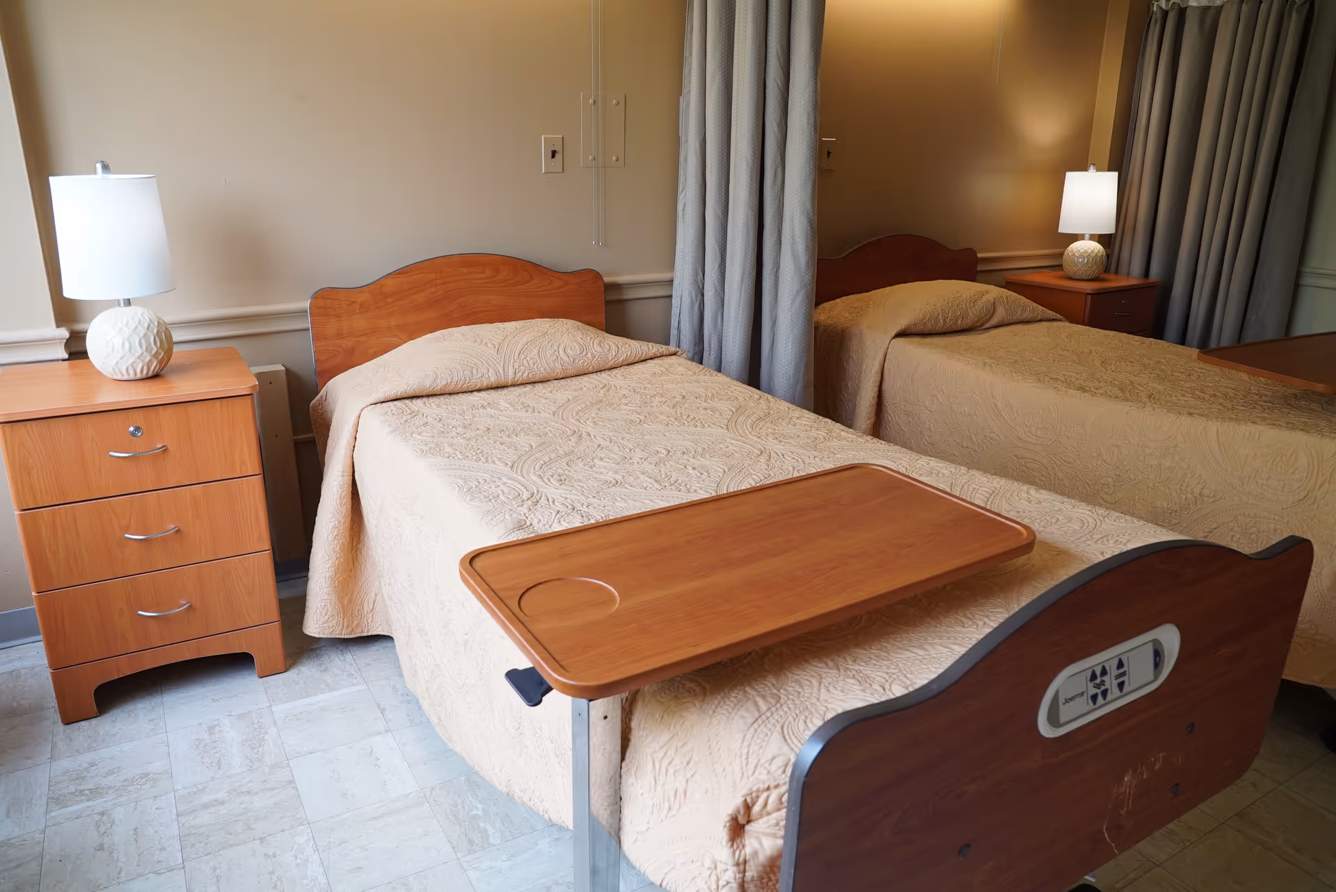 Two twin beds with wooden headboards in a senior living bedroom, each with bedside tables, lamps, and an overbed tray.