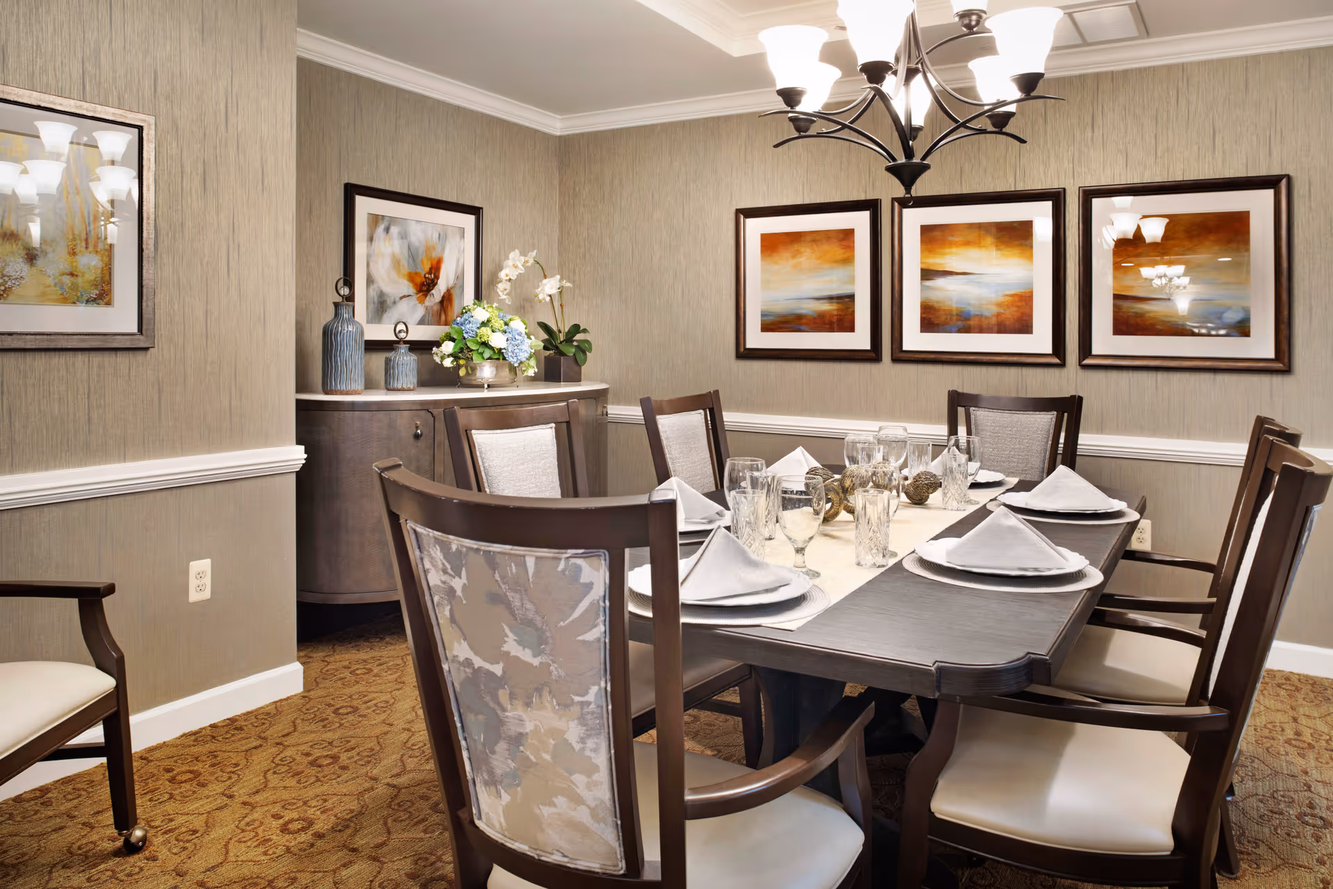 A formal dining room with a rectangular wooden table set for six people. The table is adorned with white napkins, glassware, and decorative items. The room features beige textured walls with three framed abstract paintings, a sideboard with vases and flowers, a patterned carpet, and a chandelier overhead.