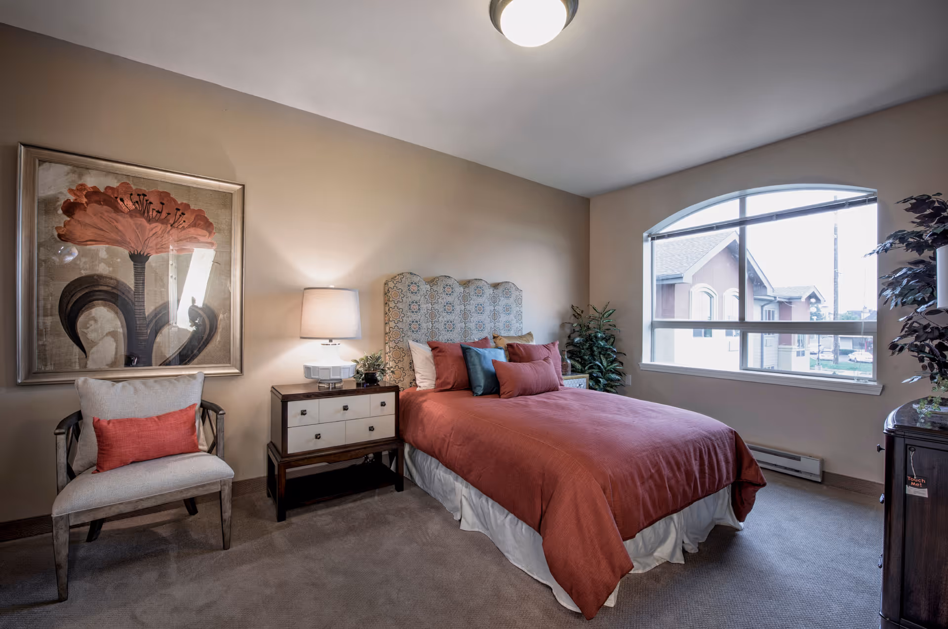 Furnished bedroom with a bed dressed in rust-colored bedding, a nightstand and lamp, an armchair beneath a large floral painting, and an arched window.