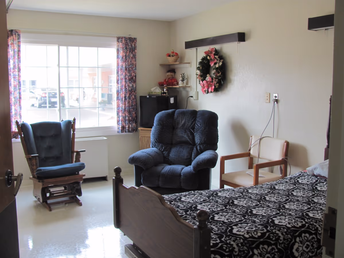 A senior living facility room with a single bed covered in a black and white patterned blanket, a blue cushioned armchair, a wooden rocking chair with blue cushions, and a beige chair. There is a large window with floral curtains letting in natural light, a small TV on a wooden stand, and a decorative wreath with pink flowers hanging on the wall.