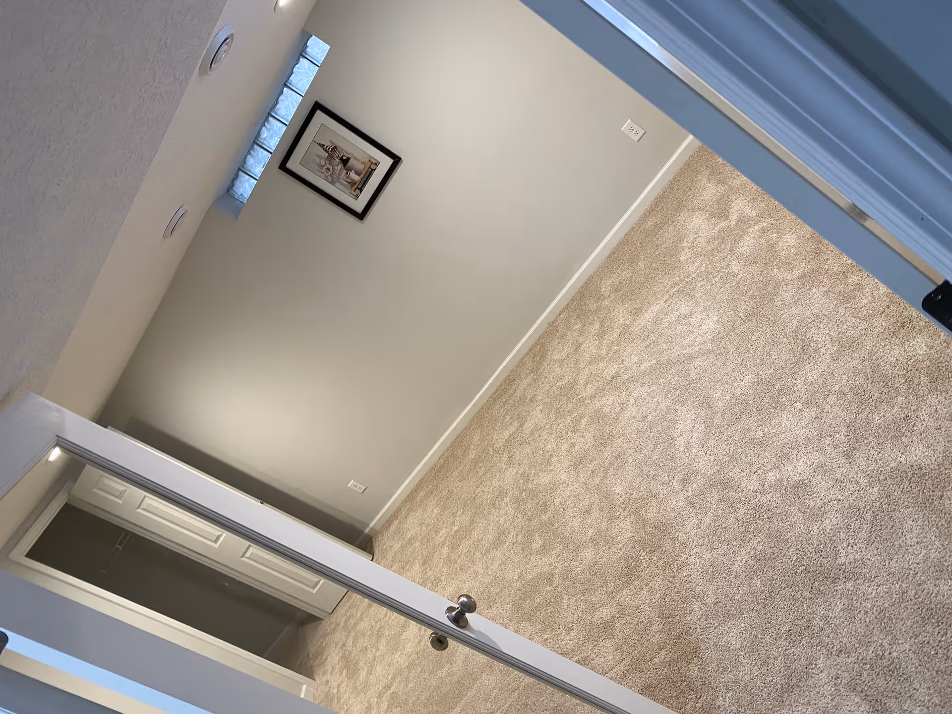 Empty carpeted bedroom with light gray walls, a framed picture, closet with bi-fold doors, and a small glass-block window.