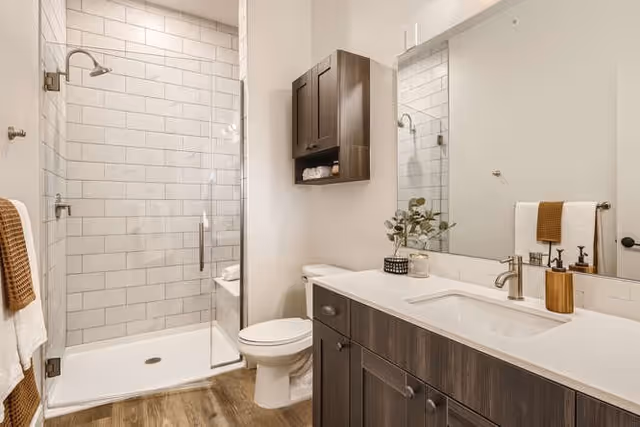 A modern bathroom featuring a glass-enclosed shower with white subway tiles, a white toilet, a wooden vanity with a white countertop and built-in sink, a large mirror above the vanity, a wall-mounted cabinet, and neatly arranged towels and decorative items.