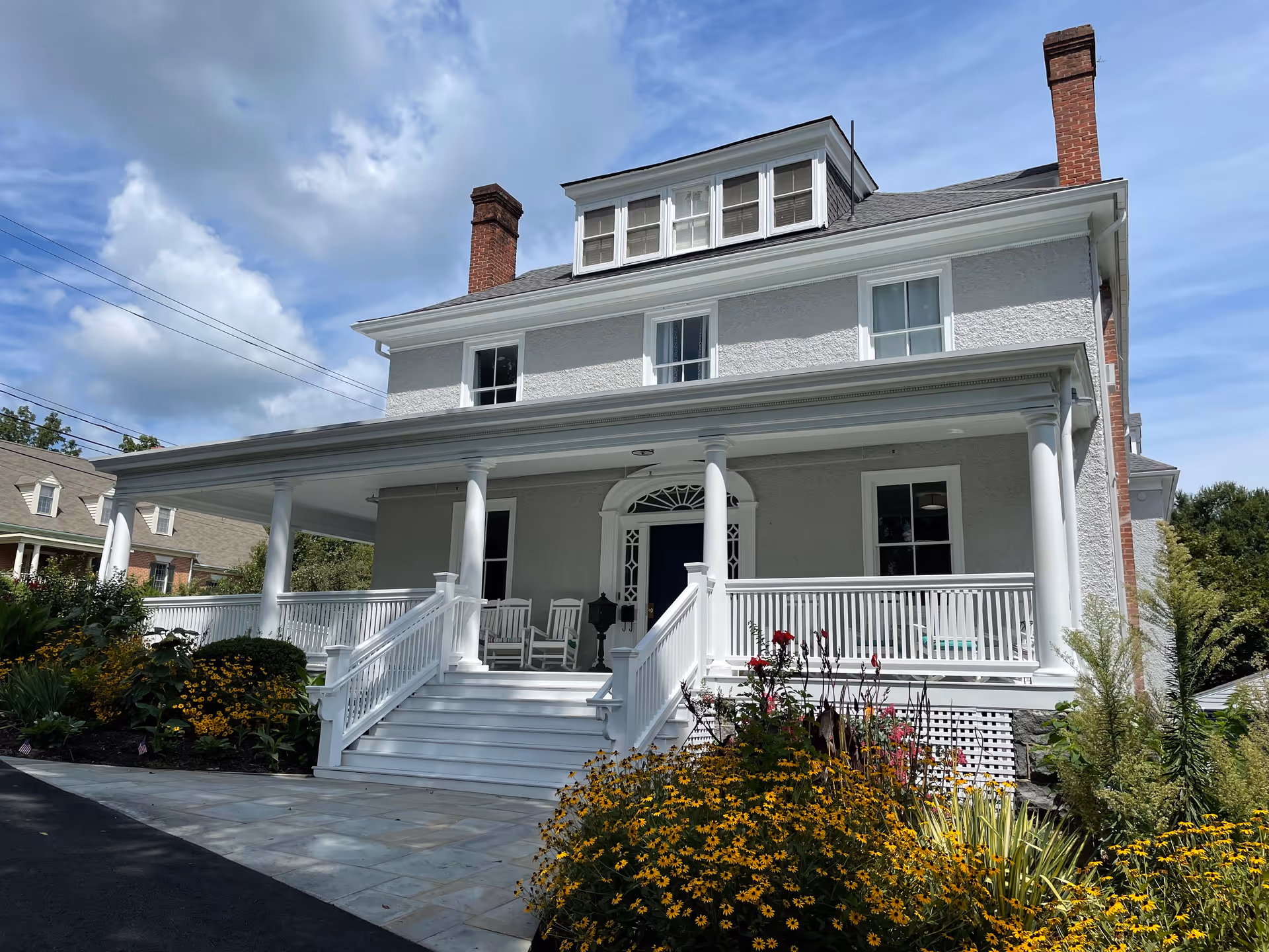 Front exterior view of a large, two-story senior living community building with a wide white porch, white railings, and steps leading up to a dark blue front door. The building has multiple windows, two brick chimneys, and is surrounded by a garden with yellow and red flowers under a partly cloudy sky.