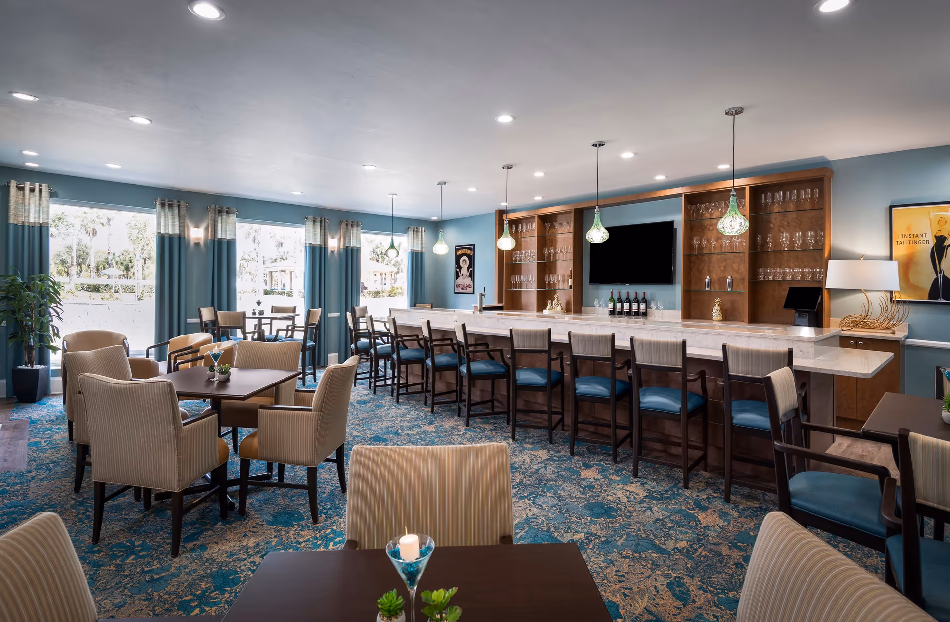 A spacious and well-lit dining area with multiple tables and chairs arranged for seating. The room features a long bar counter with high chairs, pendant lights hanging above, and shelves with glassware and bottles behind the bar. Large windows with blue curtains allow natural light to fill the space, and the carpet has a blue and beige pattern. There is a TV mounted on the wall behind the bar and framed artwork on the walls.