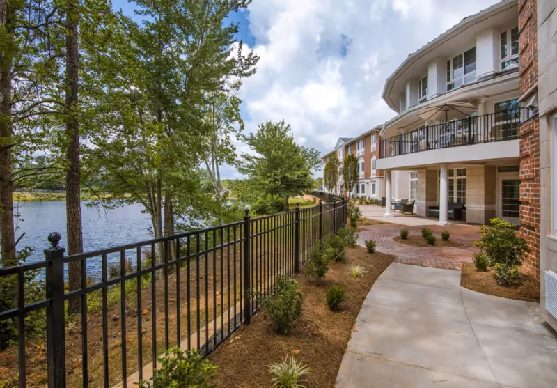 Outdoor view of The Georgian Lakeside facility showing a paved walkway alongside a black metal fence with small bushes and trees. The building has multiple floors with balconies and large windows. A body of water and more trees are visible on the left side under a partly cloudy sky.