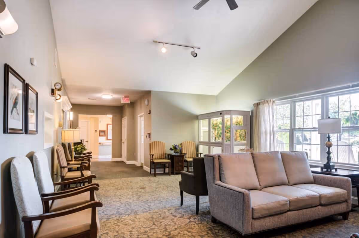 A well-lit common area with a beige sofa, several wooden chairs with cushions, a side table with a lamp, large windows with curtains, and framed bird artwork on the wall. The room has a carpeted floor and light-colored walls, with a ceiling fan and track lighting.