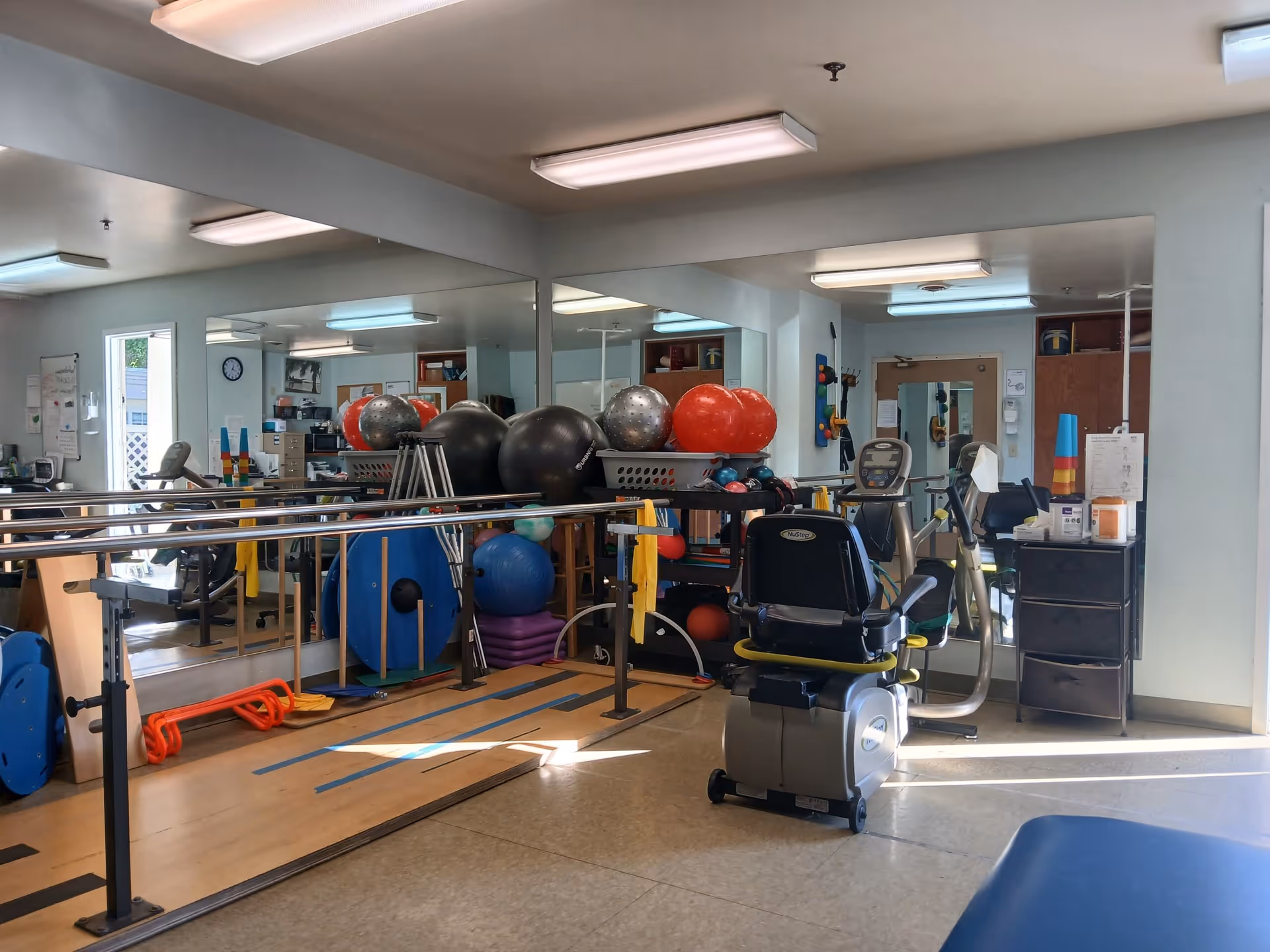 A rehabilitation or physical therapy room with exercise equipment including stationary bikes, balance balls, resistance bands, and parallel bars. The room has large mirrors on the walls and is well-lit with fluorescent ceiling lights.