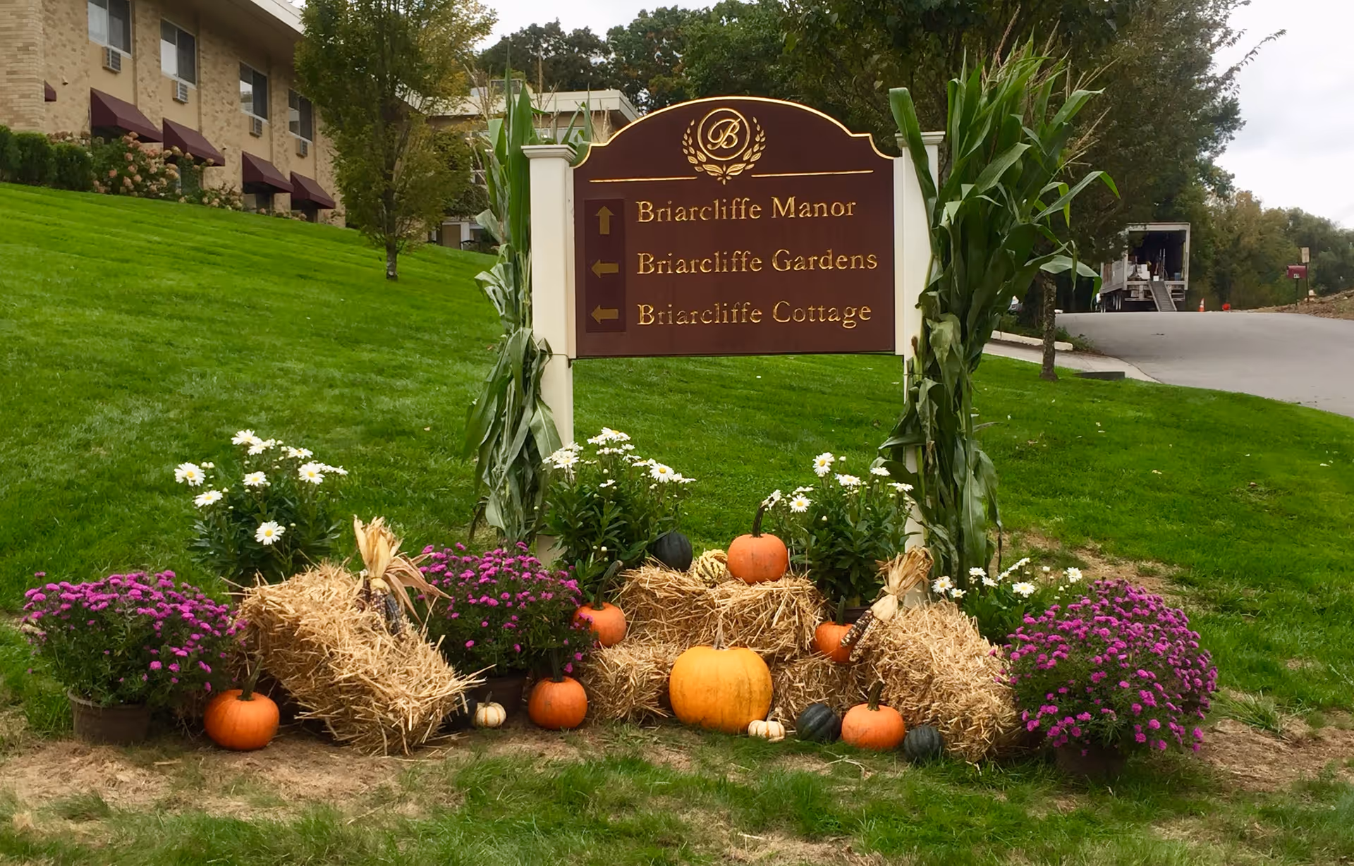 A brown and gold directional sign for Briarcliffe Manor, Briarcliffe Gardens, and Briarcliffe Cottage surrounded by fall decorations including pumpkins, hay bales, purple and white flowers, and tall corn stalks. The sign is situated on a grassy area near a building with maroon awnings and a driveway.