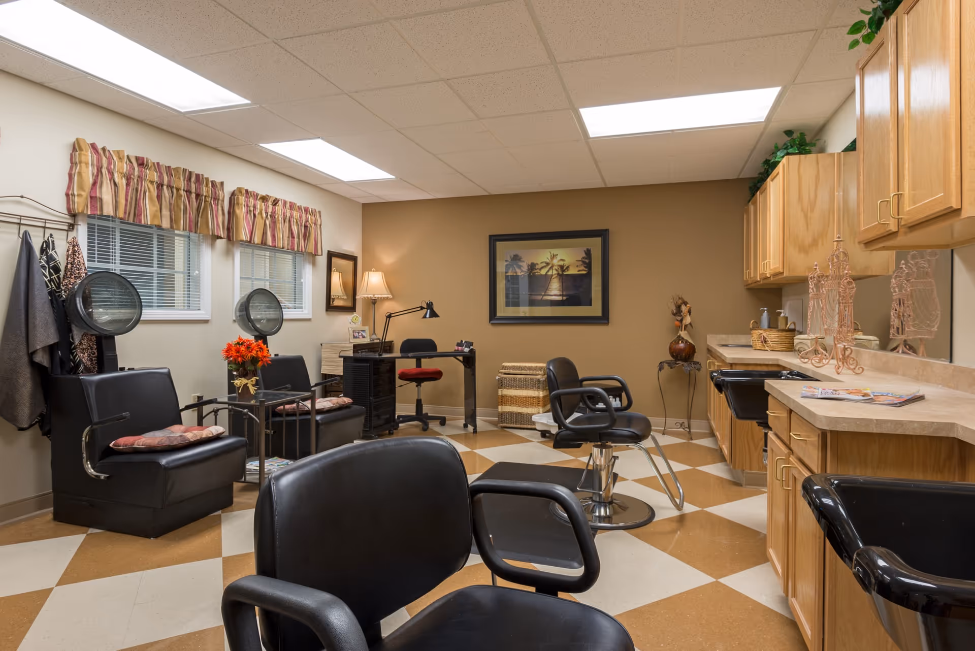 Interior of a senior living facility hair salon with black salon chairs, hair dryers, a desk with a red swivel chair, wooden cabinets, and a framed picture on a beige wall. The floor has a checkered pattern in brown and cream colors, and there are two windows with striped valances.