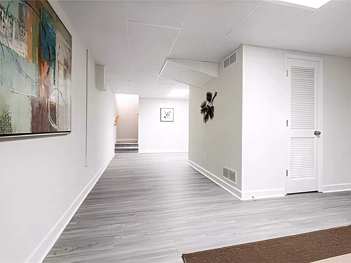 Bright, minimalist hallway with light gray wood-look flooring, white walls, wall art, and a closed louvered door.