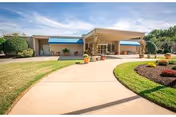 Front entrance of a single-story nursing facility with a curved driveway, landscaped lawn, potted plants, and blue awnings.
