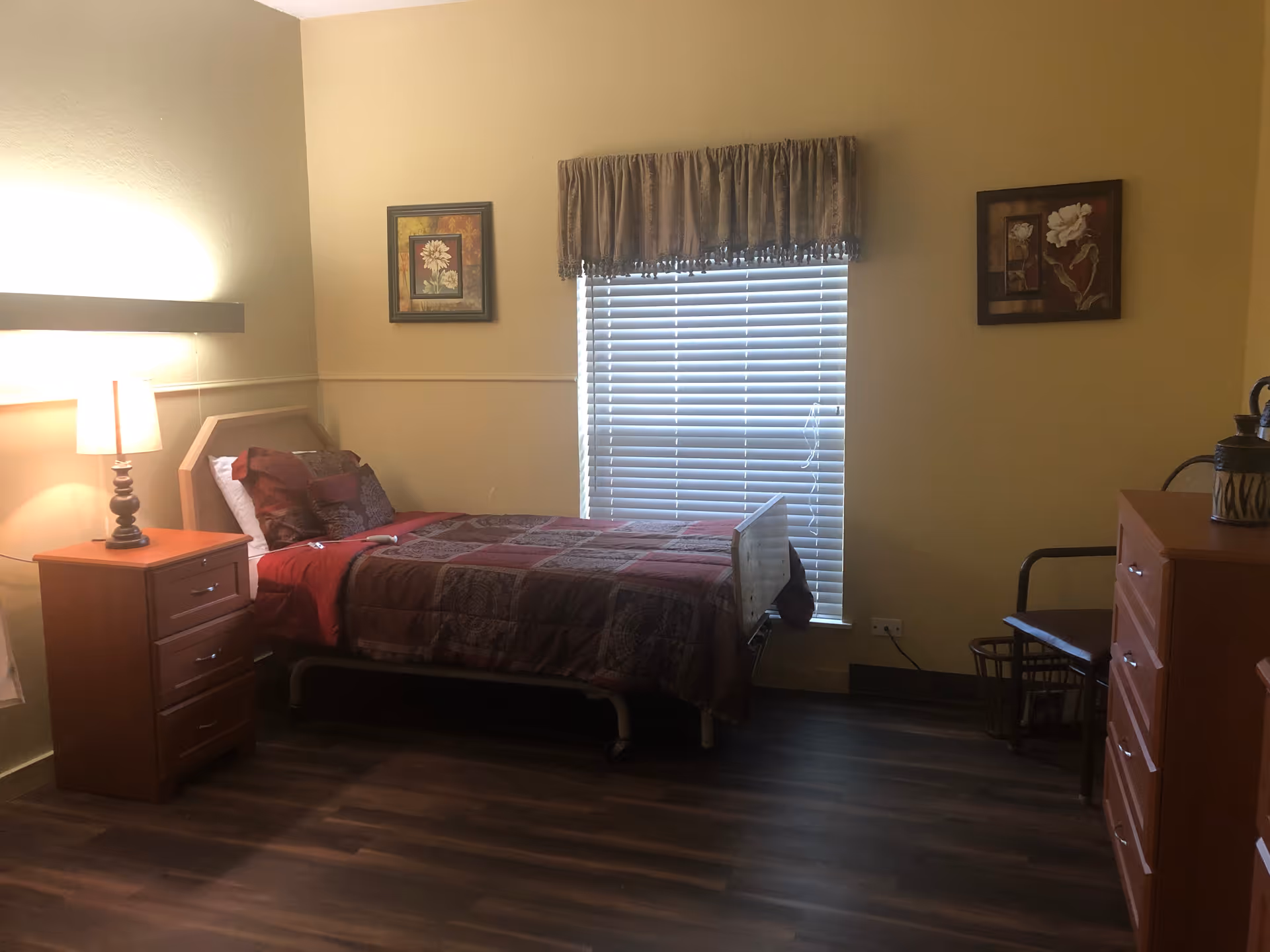 A bedroom in a senior living facility with a single bed covered in a patterned quilt and pillows. There is a wooden nightstand with a lamp on the left side of the bed. A window with closed blinds and a valance is centered on the wall behind the bed. Two framed floral pictures hang on the walls on either side of the window. On the right side of the room, there is a wooden dresser with a water bottle and a chair next to it. The floor is dark wood.