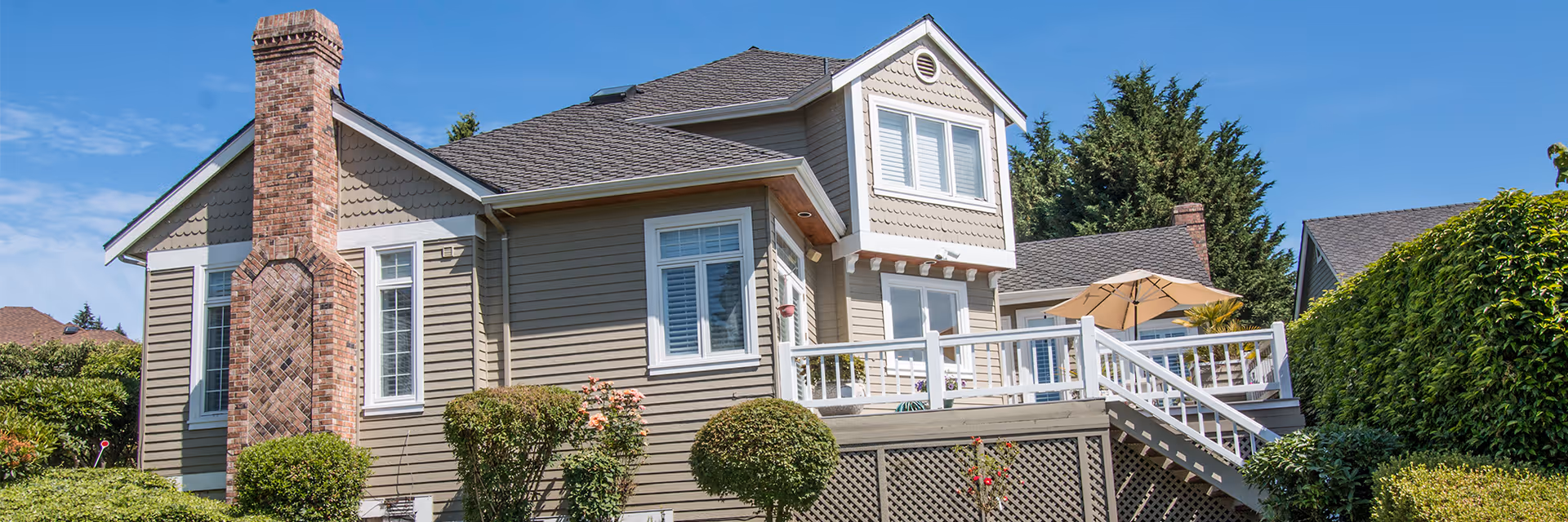 Exterior front of a beige house with a brick chimney, raised deck and patio umbrella, and landscaped shrubs under a blue sky.