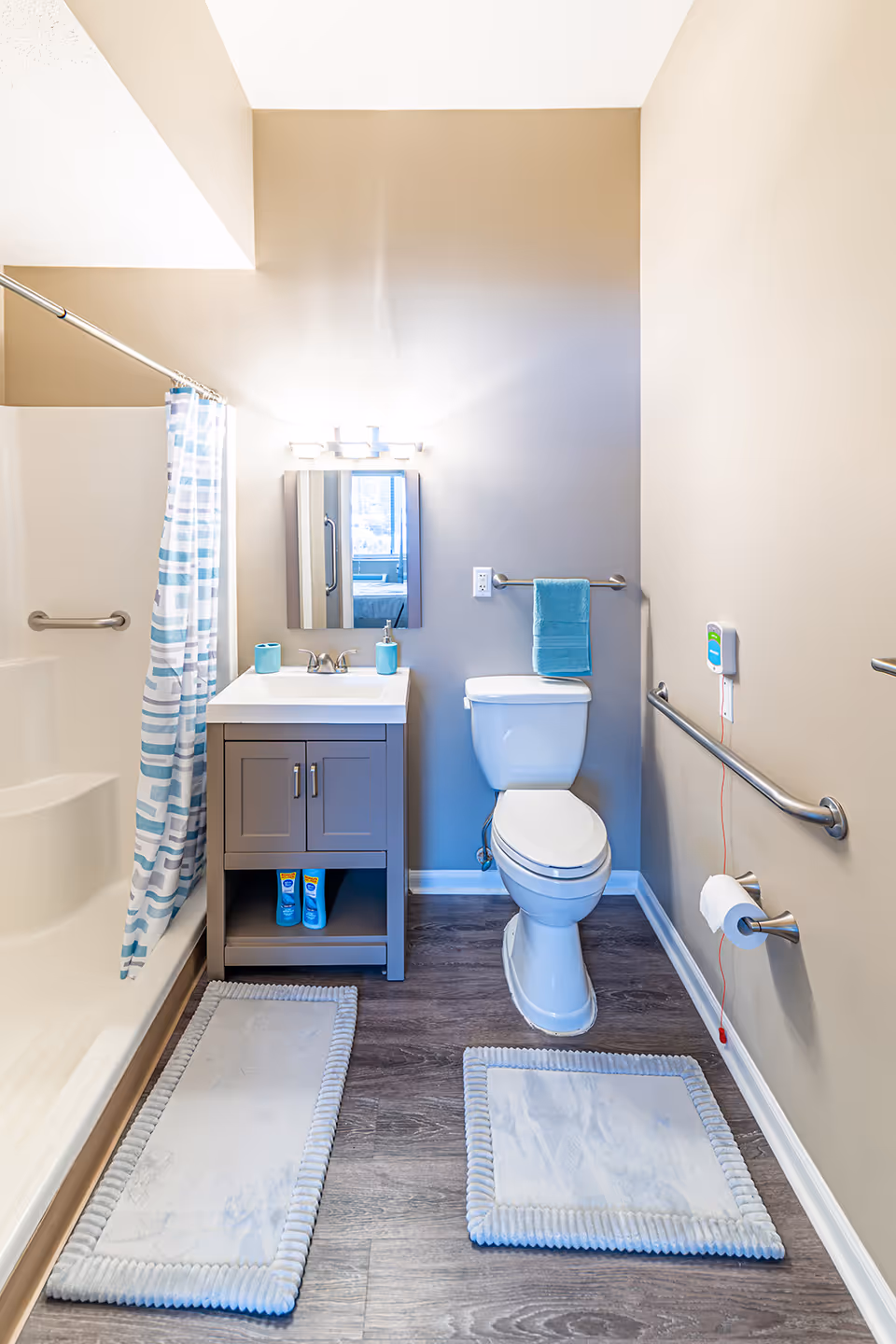 A clean and accessible bathroom featuring a white toilet with a blue towel hanging on a bar above it, a gray vanity with a white sink and mirror above, a shower with a blue and white patterned curtain, and two light gray bath mats on a wood-look floor. Grab bars are installed near the toilet and shower for safety, and an emergency pull cord is visible next to the toilet.