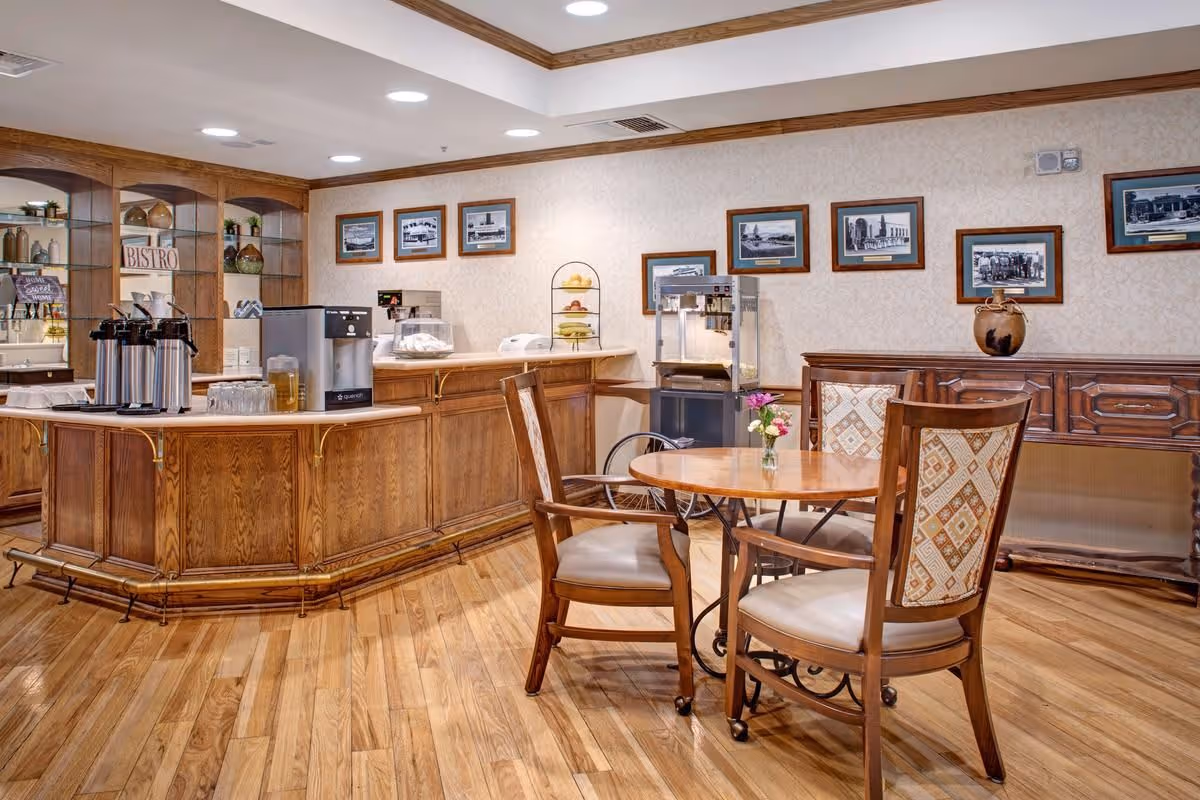 A cozy dining area in a senior living facility featuring a round wooden table with four cushioned chairs. The room has wooden flooring and a wooden counter with coffee dispensers, a water dispenser, and a popcorn machine. The walls are decorated with framed black and white photographs and a small vase with flowers is placed on the table.