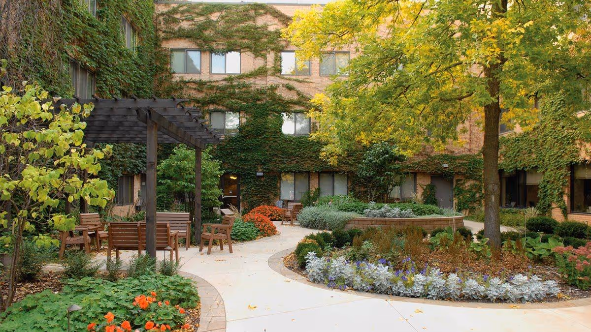 A peaceful outdoor garden area at Jones-Harrison Senior Living featuring a curved concrete pathway, wooden benches under a pergola, various green plants, colorful flowers, and a large tree. The building walls are covered with ivy.