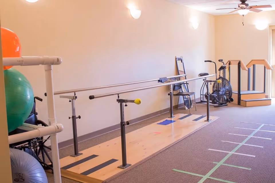 A physical therapy room with parallel bars for walking exercises, exercise balls, a stationary bike, a chair, and a small set of stairs with handrails. The room has beige walls, carpeted floor, and ceiling lights.